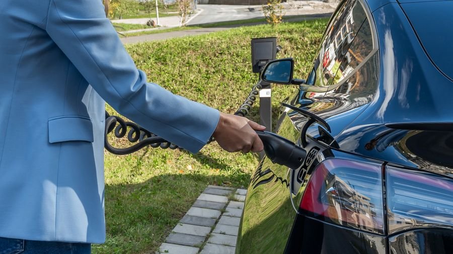 Mujer con chaqueta azul conectando el cable de carga a un carro eléctrico oscuro junto a un prado