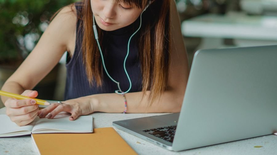 Mujer con audífonos toma notas en un cuaderno frente a un portátil durante una clase en línea
