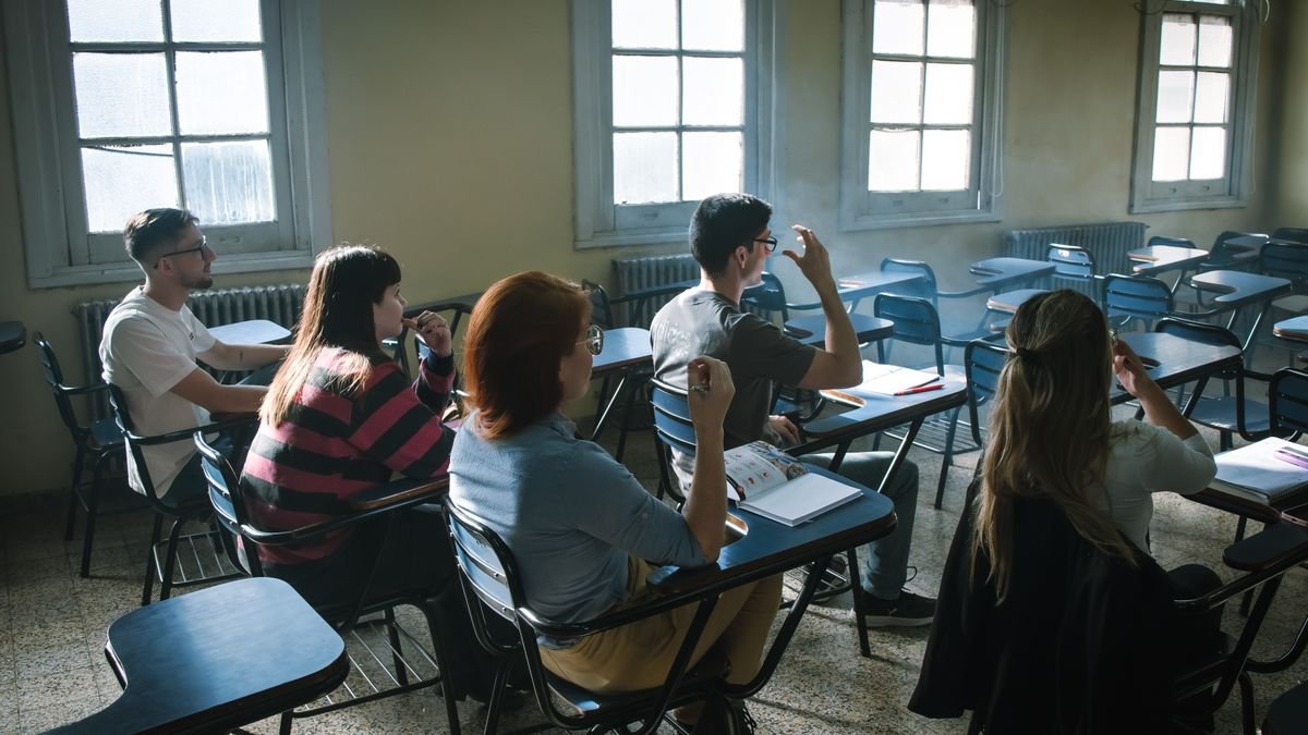 Adultos sentados en pupitres de un salón de clase tomando apuntes mientras uno levanta la mano